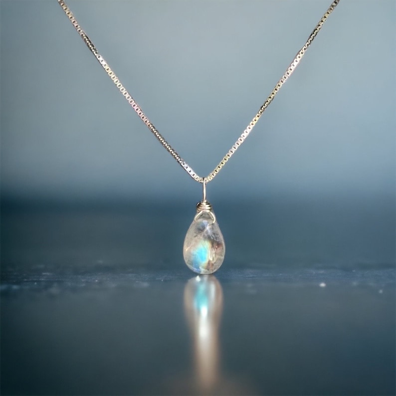 May include: A silver necklace with a teardrop-shaped pendant. The pendant is a clear stone with blue and white internal colors. The necklace chain is a delicate, square-link style, and the pendant is wire-wrapped.