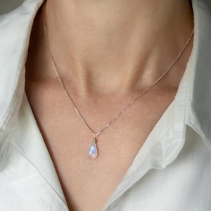 May include: A delicate silver necklace with a teardrop-shaped moonstone pendant. The moonstone displays a soft, iridescent glow, suspended from a fine chain. The necklace is worn against a white collared shirt.