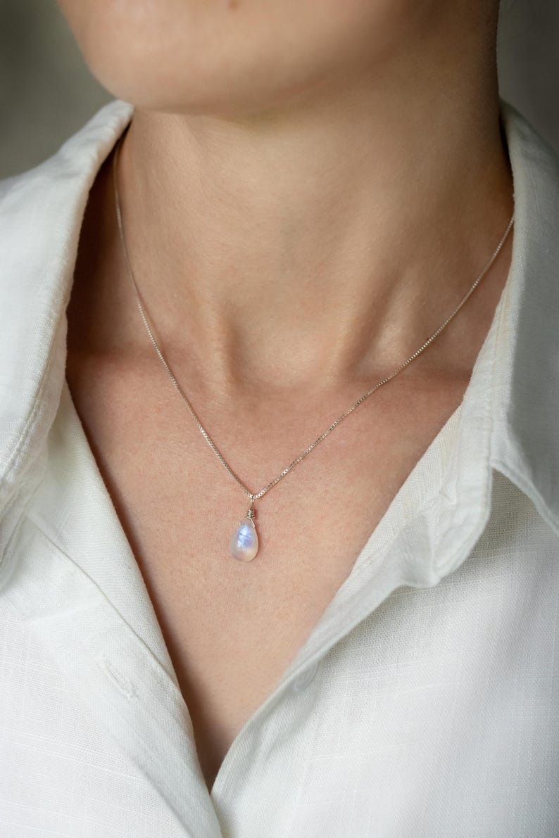 May include: A delicate silver necklace with a teardrop-shaped moonstone pendant. The moonstone displays a soft, iridescent glow, suspended from a fine chain. The necklace is worn against a white collared shirt.