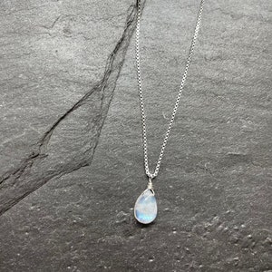 May include: A silver necklace with a pear-shaped, iridescent moonstone pendant. The pendant is wire-wrapped and hangs from a delicate chain. The necklace rests on a dark gray, textured surface.