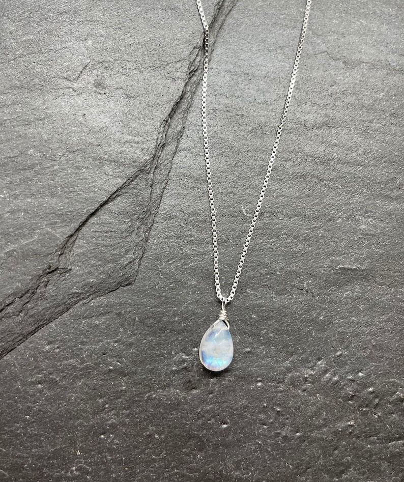 May include: A silver necklace with a pear-shaped, iridescent moonstone pendant. The pendant is wire-wrapped and hangs from a delicate chain. The necklace rests on a dark gray, textured surface.