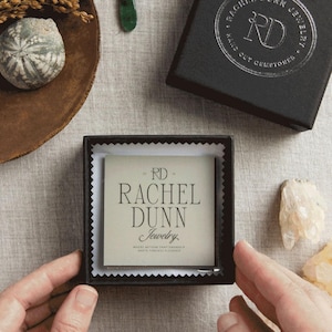 May include: An overhead shot of jewelry and packaging. The image features a pair of green teardrop earrings, a black jewelry box with silver text, and a smaller box with the text "RACHEL DUNN Jewelry". The background is a neutral linen fabric.