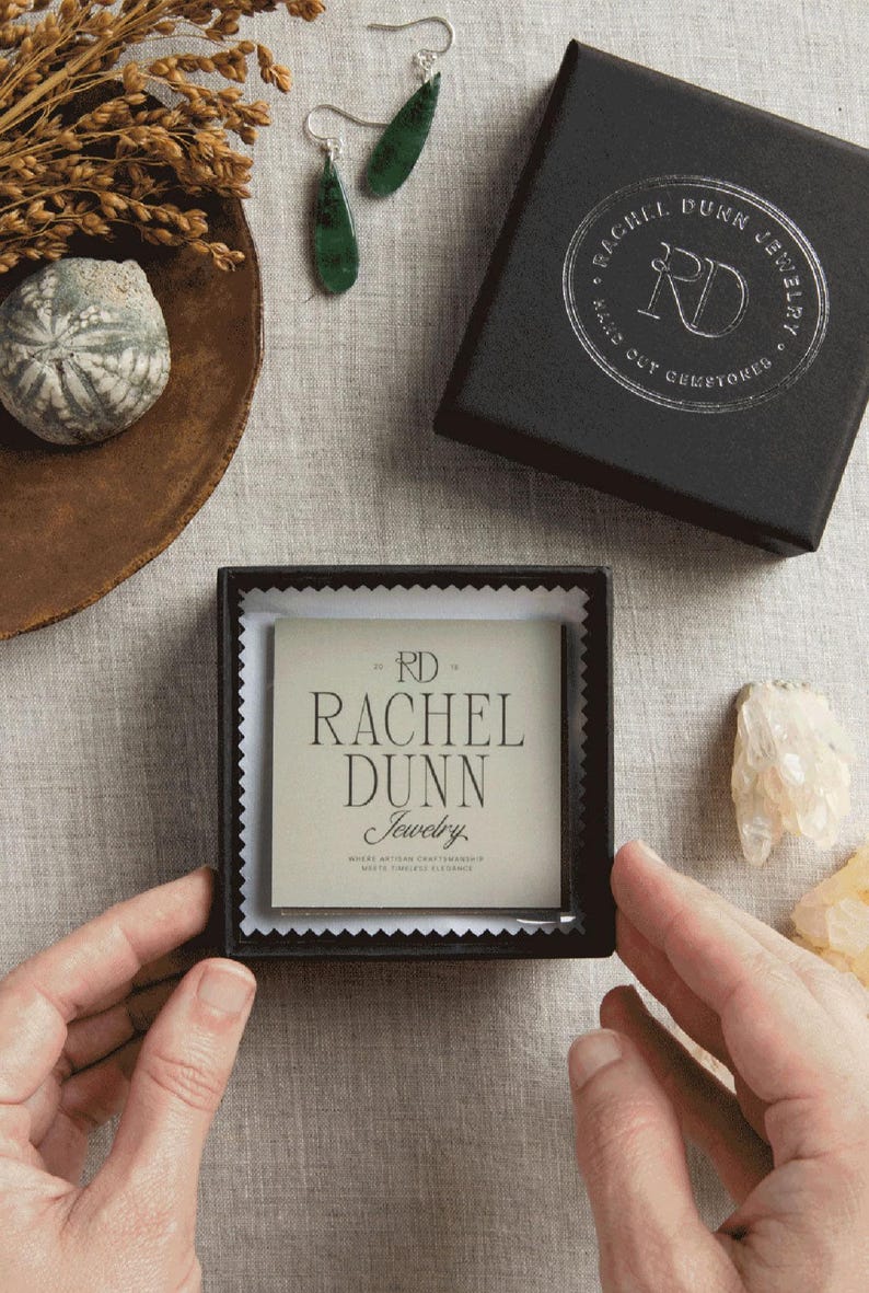 May include: An overhead shot of jewelry and packaging. The image features a pair of green teardrop earrings, a black jewelry box with silver text, and a smaller box with the text "RACHEL DUNN Jewelry". The background is a neutral linen fabric.