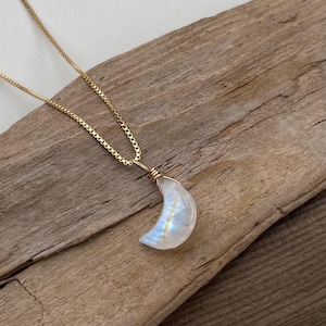 Könnte beinhalten: Eine goldene Halskette mit einem Mond-Anhänger. Der Mond-Anhänger ist hellblau und weiß, mit einer Golddrahtwicklung. Die Halskette wird auf einem Stück verwittertem Holz präsentiert.