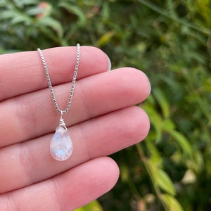 May include: A delicate silver necklace featuring a pear-shaped, iridescent moonstone pendant. The pendant is wire-wrapped and suspended from a fine silver chain. The necklace is displayed against a blurred green background.