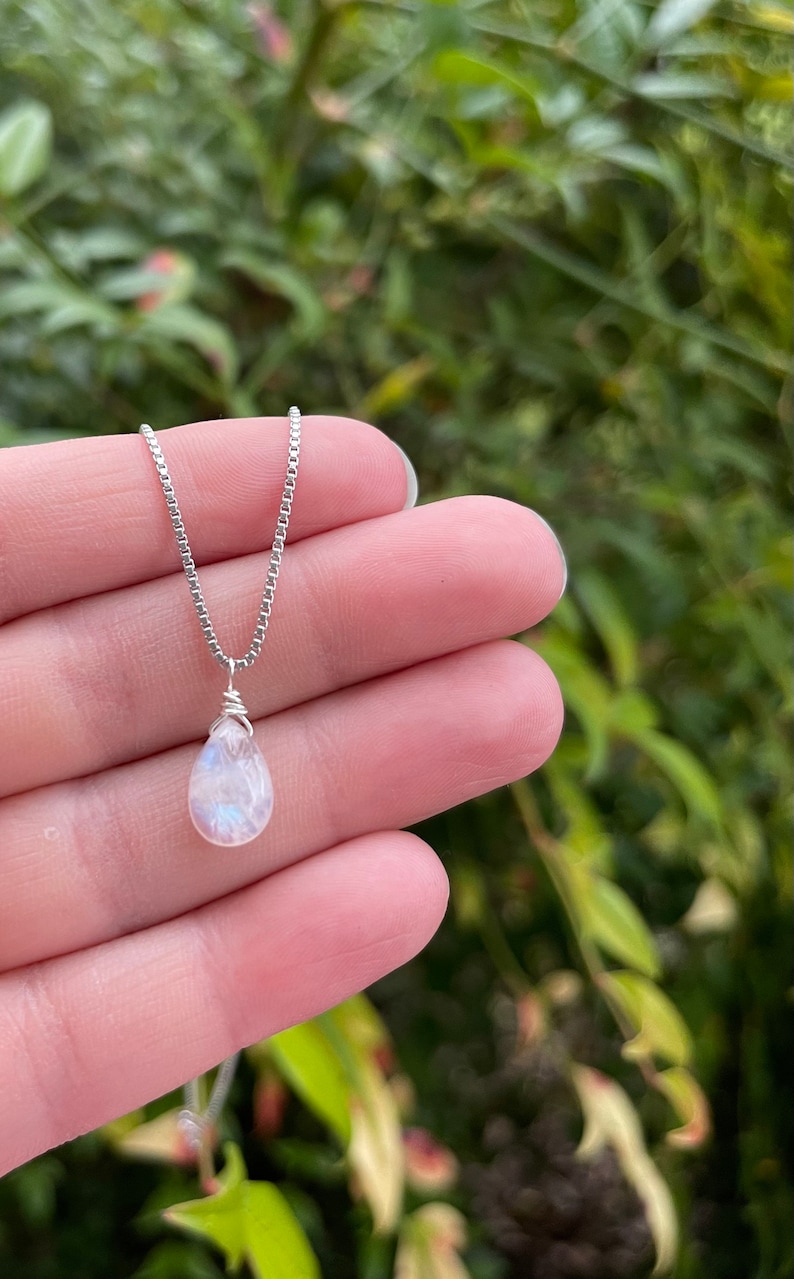 May include: A delicate silver necklace featuring a pear-shaped, iridescent moonstone pendant. The pendant is wire-wrapped and suspended from a fine silver chain. The necklace is displayed against a blurred green background.