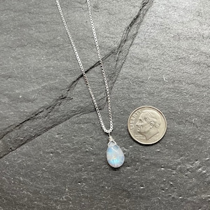 May include: A silver necklace with a pear-shaped, iridescent gemstone pendant. The pendant is wire-wrapped and hangs from a delicate chain. A U.S. dime is next to the necklace for size comparison. The necklace is displayed on a dark gray surface.