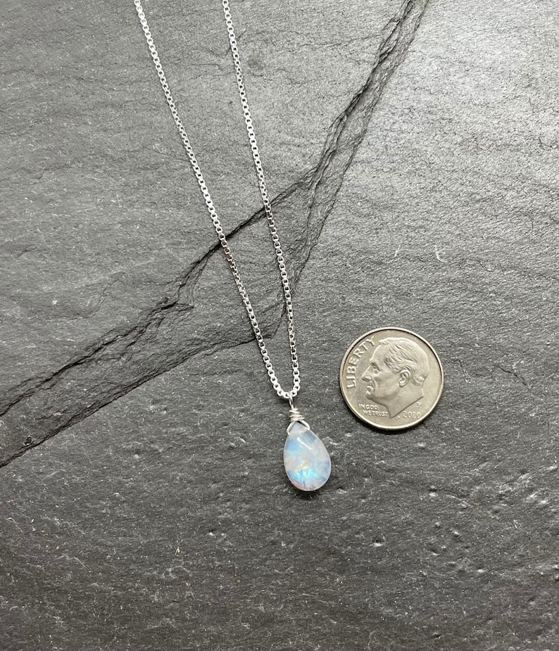 May include: A silver necklace with a pear-shaped, iridescent gemstone pendant. The pendant is wire-wrapped and hangs from a delicate chain. A U.S. dime is next to the necklace for size comparison. The necklace is displayed on a dark gray surface.