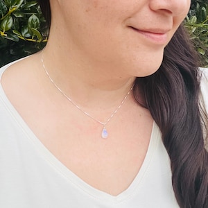 May include: A delicate silver necklace featuring a small, pear-shaped moonstone pendant. The pendant displays a subtle play of colors, with hints of blue and white. The necklace is a simple, elegant piece of jewelry.
