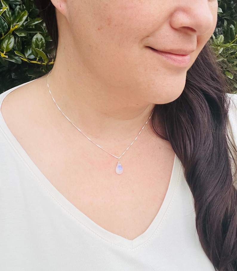 May include: A delicate silver necklace featuring a small, pear-shaped moonstone pendant. The pendant displays a subtle play of colors, with hints of blue and white. The necklace is a simple, elegant piece of jewelry.