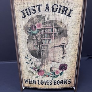 May include: A book folding art piece with a brown cover. The front cover features a woman's face with a bookshelf for hair. The text reads "Just a girl who loves books".