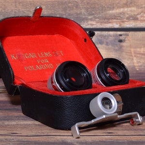 May include: A black leather case with a red interior, containing two black camera lenses and a silver lens attachment. The case has a gold inscription that reads "KAGAR LENS SET FOR POLAROID".