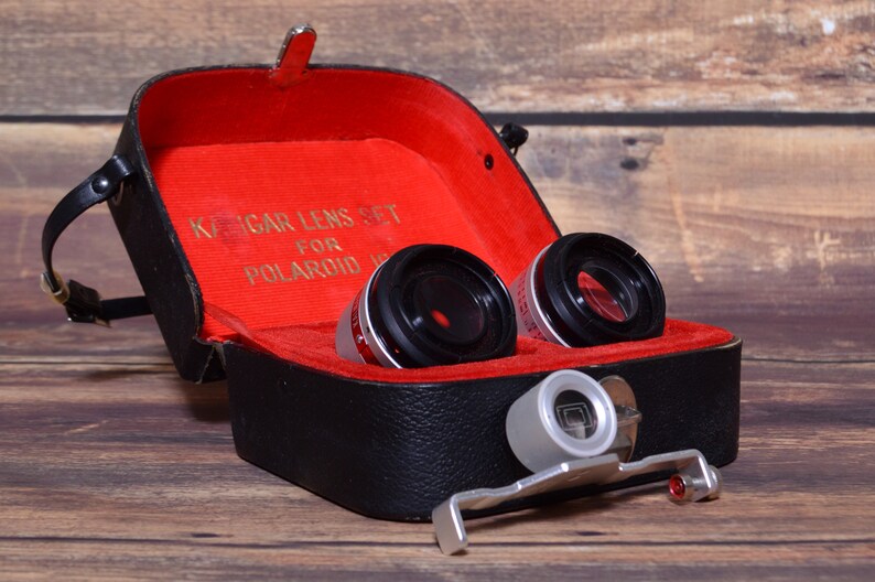 May include: A black leather case with a red interior, containing two black camera lenses and a silver lens attachment. The case has a gold inscription that reads "KAGAR LENS SET FOR POLAROID".
