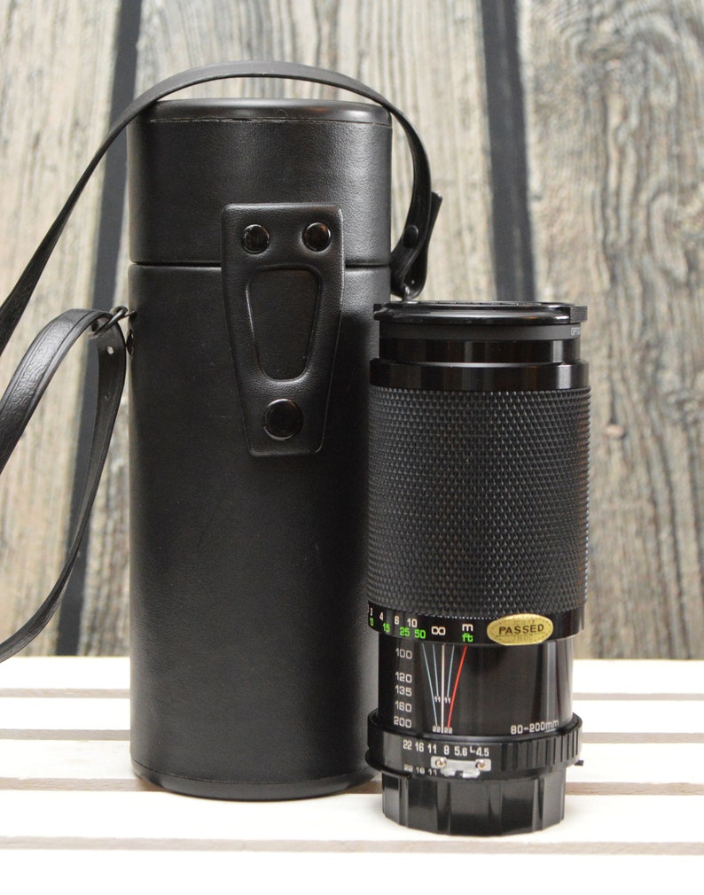 May include: A black camera lens with a black leather carrying case. The lens has a zoom range of 80-200mm and a maximum aperture of f/5.6-4.5. The lens has a depth of field scale and a focus ring. The lens is in good condition.