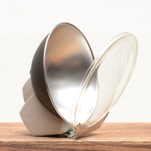 May include: A studio photography light modifier, featuring a silver reflector, a black dome, and a clear plastic diffuser. The light modifier is mounted on a white and gray base, designed to direct and soften light for professional photography.