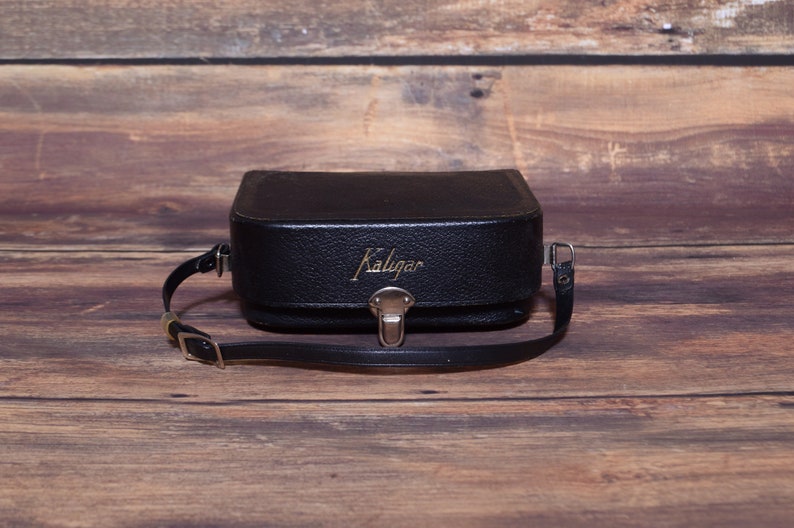 May include: A black leather camera case with a gold Kaligar logo and a black adjustable strap.