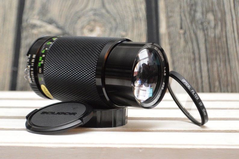 May include: A black camera lens with a 55-200mm zoom range and a 1:4.5 aperture. The lens has a black lens cap and a black filter attached to the front.