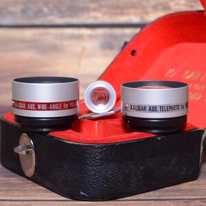May include: Two silver camera lens attachments in a black leather case with a red velvet lining. The lens on the left says "KALIGAR AUX. WIDE-ANGLE for POLA" and the lens on the right says "KALIGAR AUX. TELEPHOTO for 16MM".