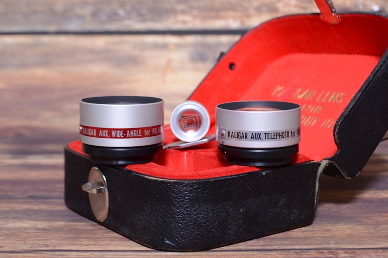 May include: Two silver camera lens attachments in a black leather case with a red velvet lining. The lens on the left says "KALIGAR AUX. WIDE-ANGLE for POLA" and the lens on the right says "KALIGAR AUX. TELEPHOTO for 16MM".