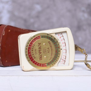 May include: A white and gold Ikophot light meter with a brown leather case and a gold chain. The meter has a dial with markings for ASA and DIN film speeds.