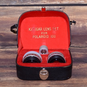May include: A black leather case with a red interior, containing a set of three silver camera lenses. The case has a label that reads "Kaigar Lens Set for Polaroid 100".