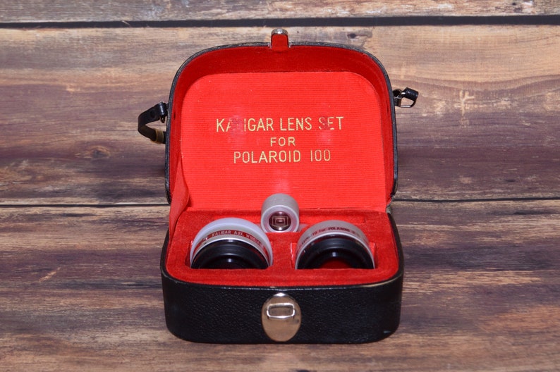 May include: A black leather case with a red interior, containing a set of three silver camera lenses. The case has a label that reads "Kaigar Lens Set for Polaroid 100".