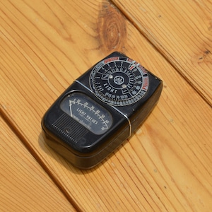 May include: A black vintage General Electric light meter with a circular dial and a needle. The dial has markings for light values and shutter speeds. The meter has a black plastic case with a ribbed texture.
