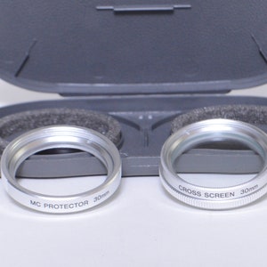 May include: Two silver camera lens filters in a gray case. The filter on the left says "MC PROTECTOR 30mm" and the filter on the right says "CROSS SCREEN 30mm".