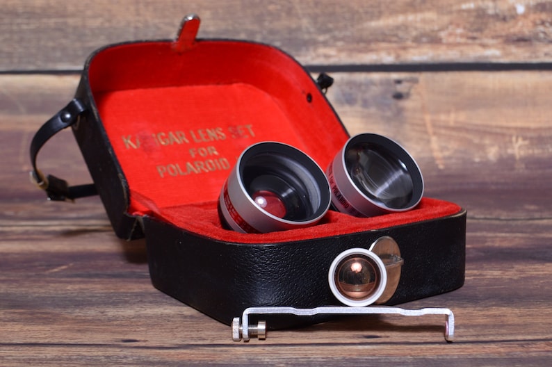 May include: A black leather case with a red interior holds two silver and black camera lenses. The case has a gold inscription that reads "Kodak Lens Set for Polaroid".