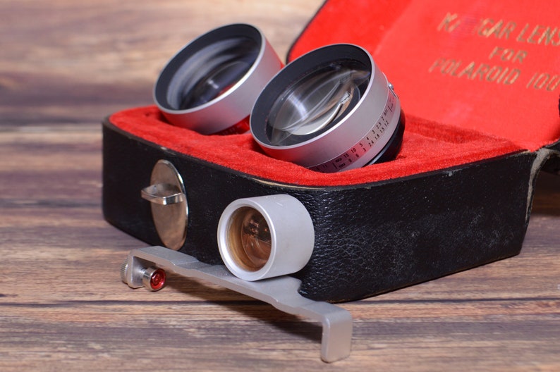 May include: A set of three silver camera lenses in a black and red leather case. The lenses are labeled "Carl Zeiss" and "Polaroid 100".