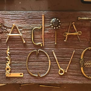 May include: A rectangular piece of weathered, dark brown wood features the words RADIATE LOVE spelled out using various brass-toned metal hardware components, including hooks, brackets, and fasteners, accented by small red floral embellishments.