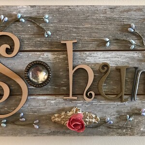 May include: A rectangular wall sign made of three horizontal weathered gray wooden planks. The word Sohum is displayed with a large wooden S, a circular crystal knob as the o, a wooden h, a metal u, and two metal nails forming the m.