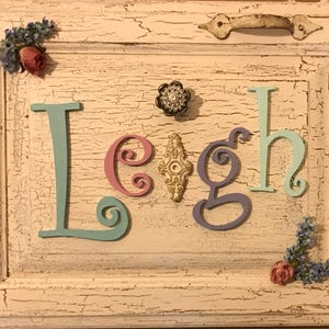 May include: A rectangular white wooden cabinet door with a distressed crackle finish features the name Leigh in colorful cutout letters. The letter L is light blue, e is pink, i is a gold ornament, g is purple, and h is light blue. Floral accents decorate the corners.