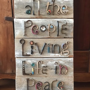 May include: A rectangular wall sign made of weathered, light gray horizontal wooden planks. The text "imagine all the People Living Life in peace" is spelled out using various metal hardware, including nails, screws, hooks, and cabinet pulls, accented with small colorful glass beads.