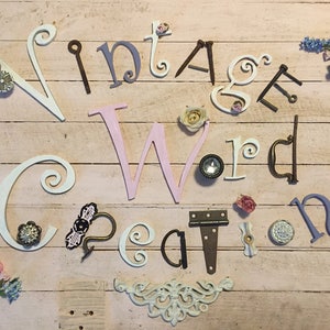 May include: The text Vintage Word Creations is spelled out using a mix of white, pink, and purple decorative letters, metal hardware pieces, and small fabric flowers arranged on a light pink wooden surface with horizontal planks.