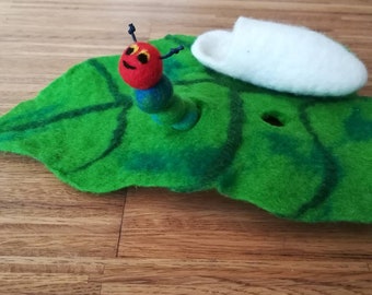 Puede incluir: Una oruga de fieltro con cabeza roja, ojos amarillos y cuerpo verde, situada sobre una gran hoja de fieltro verde con un patrón de venas verde oscuro. Un capullo de fieltro blanco descansa sobre la hoja junto a la oruga.