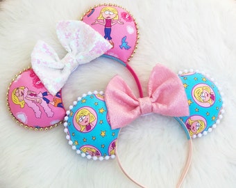 May include: A pair of pink and blue mouse ears with a white bow and a pink bow. The ears are decorated with a cartoon character and stars.