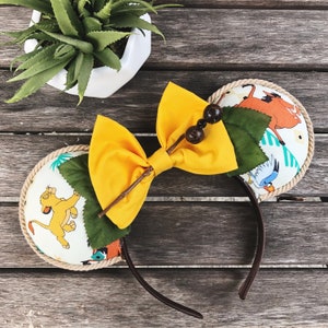 May include: A pair of mouse ears with a yellow bow and decorative accents. The ears feature a rope trim and fabric with cartoon characters. The bow is adorned with wooden beads and twigs. The headband is brown.