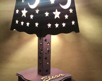 May include: A purple wooden nightlight lamp with a black shade featuring cut-out stars and moons. The lamp has a small drawer with the name "Elisa" engraved on the front.