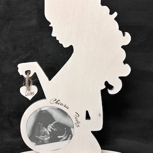 Può includere: Silhouette in legno bianco di una donna incinta che tiene un piccolo cuore con il nome "Leone" su di esso. La silhouette ha un'apertura circolare per una foto, con un'immagine di ecografia all'interno. Il testo "Chiara Fodes" è inciso sul legno.