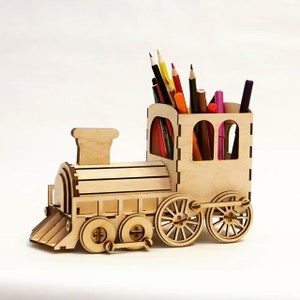 Può includere: Portapenne in legno a forma di treno giocattolo con finitura in legno marrone. Il treno ha un camino e ruote nere. Il portapenne è pieno di matite colorate e pennarelli.