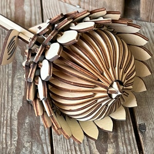 Puede incluir: Una escultura de flor de madera con detalles intrincados, hecha de piezas de madera superpuestas. La flor es marrón y beige y tiene una forma redondeada.