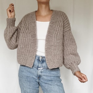 Könnte beinhalten: Eine beige Strickjacke aus grobem Strick mit gerippter Textur, langen Ärmeln und lockerer Passform. Die Strickjacke wird über einem schlichten weißen Tanktop und einer hellblauen Jeans getragen. Das Strickmuster weist durchgehend vertikale Rippen auf.