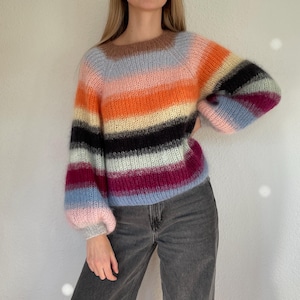 Könnte beinhalten: Ein mehrfarbiger gestreifter Pullover mit Rundhalsausschnitt und langen, puffärmeligen Ärmeln. Der Pullover hat horizontale Streifen in verschiedenen Farben, darunter Rosa, Orange, Schwarz und Blau. Das Model trägt eine dunkelgraue Jeans.