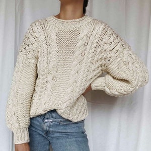 Könnte beinhalten: Ein cremefarbener Strickpullover mit Zopfmuster, lockerer Passform und langen Ärmeln. Der Pullover ist aus weichem, dickem Garn gefertigt und hat einen leicht überdimensionalen Look.