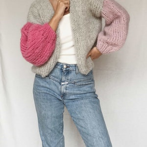 May include: A gray and pink knitted cardigan with a V-neck and long sleeves. The sleeves are puffed and the cardigan is cropped.