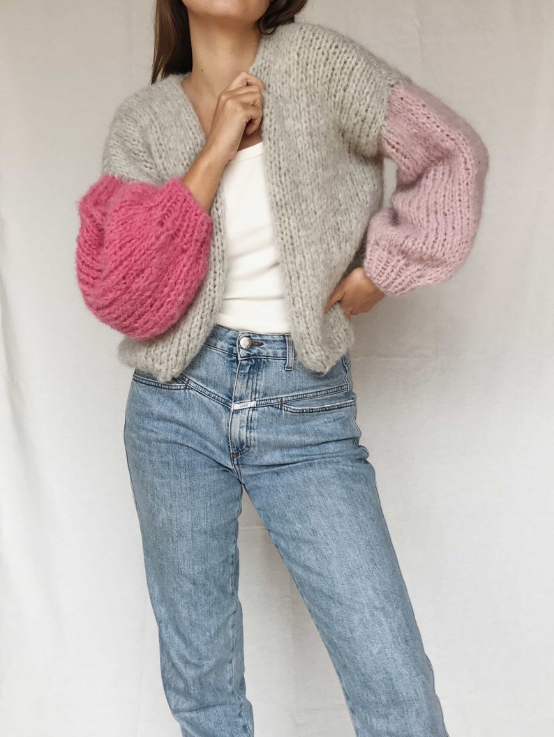 May include: A gray and pink knitted cardigan with a V-neck and long sleeves. The sleeves are puffed and the cardigan is cropped.