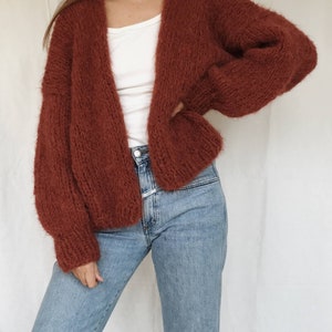 May include: A rust-colored fuzzy cardigan with long sleeves and an open front. The cardigan is worn over a white t-shirt and blue jeans.