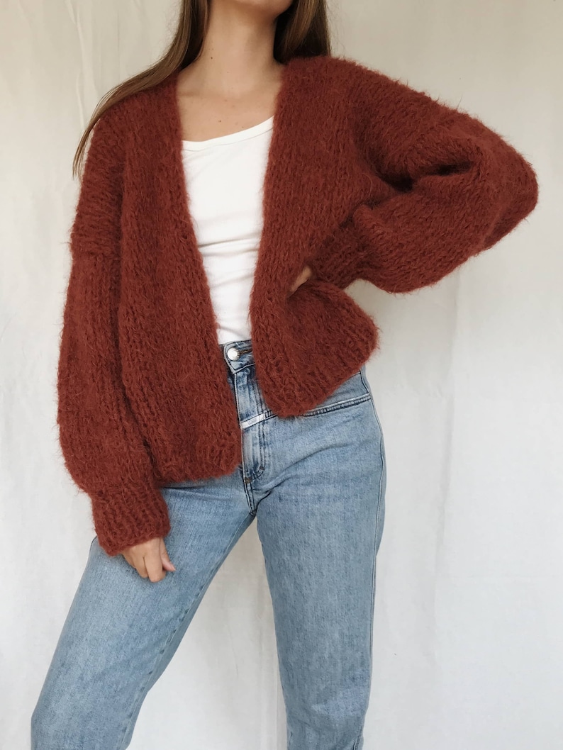 May include: A rust-colored fuzzy cardigan with long sleeves and an open front. The cardigan is worn over a white t-shirt and blue jeans.