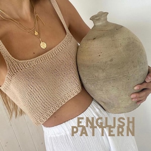 May include: A beige knitted crop top with a square neckline. The top is paired with white pants. A gold necklace with multiple layers and a pendant is visible. A large, beige, textured vase is held in the background. The text "ENGLISH PATTERN" is visible.